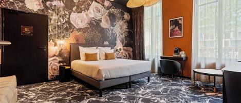 A hotel room with a large bed, a desk, and a chair. There is a door with a sign, a wall with a floral mural, and a window with curtains.