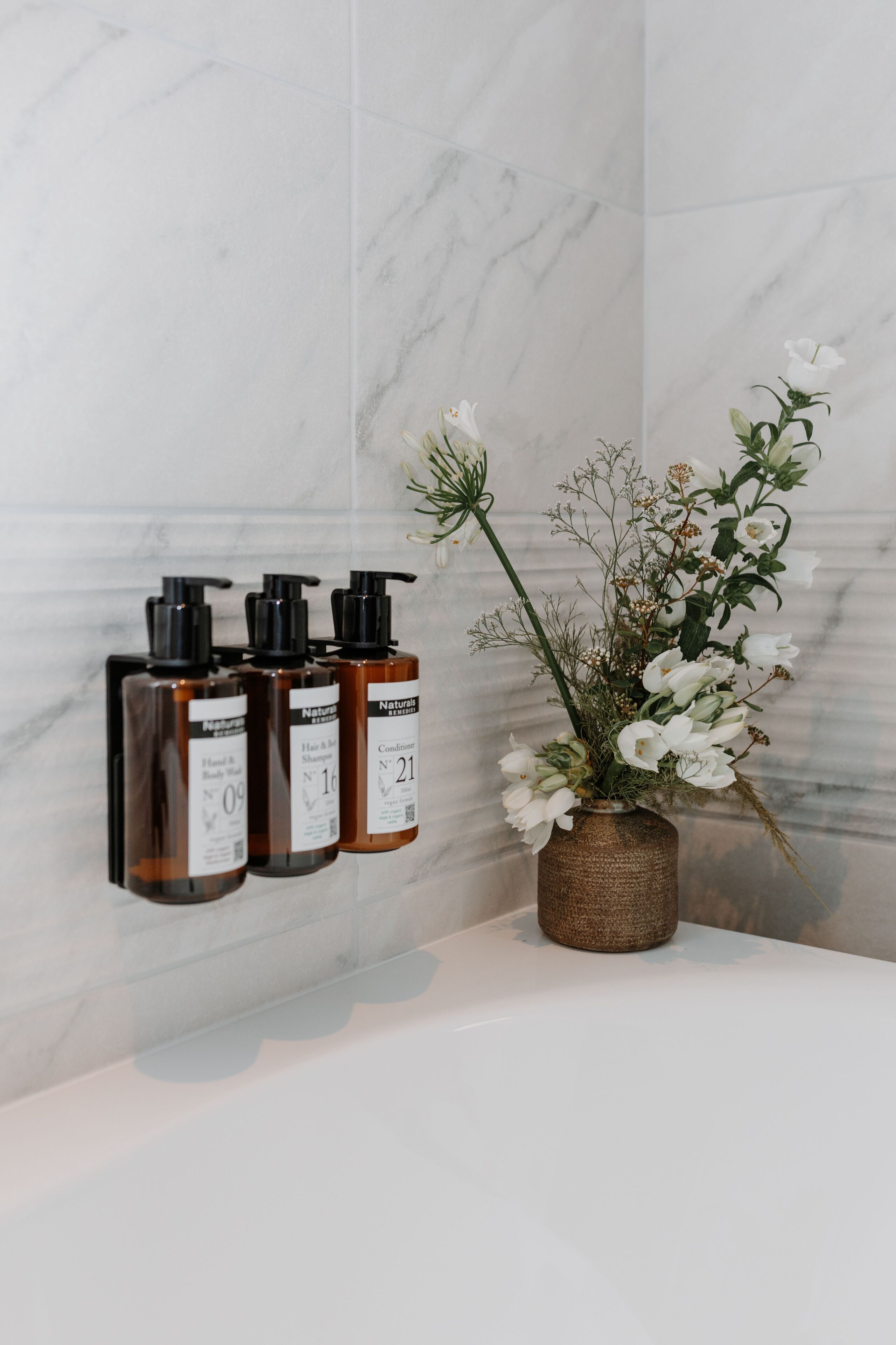A bathroom with marble walls, a white bathtub, and three brown bottles labeled "Natural" on a shelf.