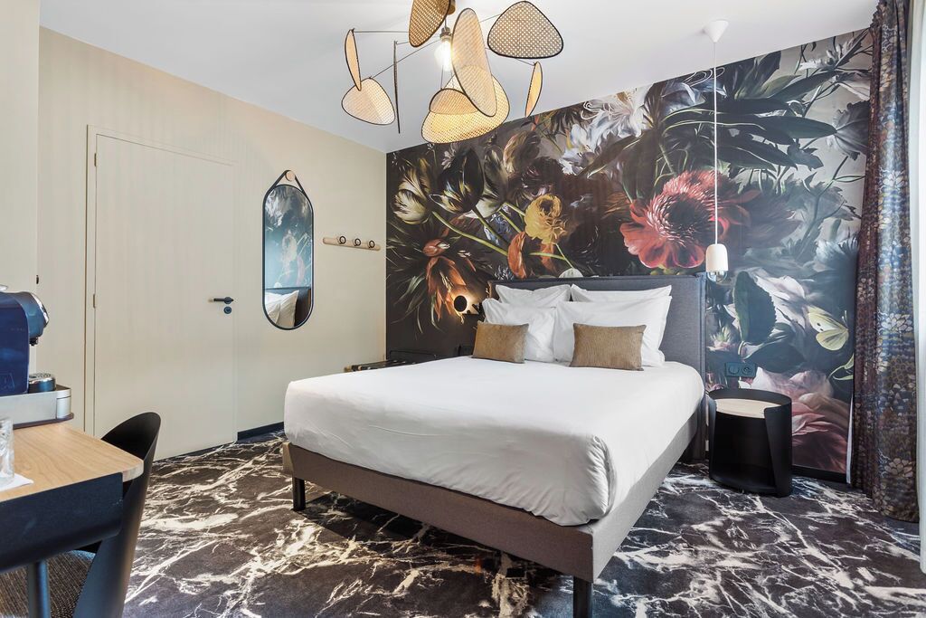 A hotel room with a bed, a desk, and a chair. There is a large floral mural on the wall and a ceiling light fixture.