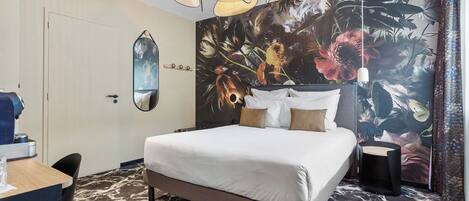 A hotel room with a bed, a desk, and a chair. There is a large floral mural on the wall and a ceiling light fixture.