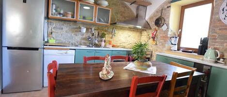 A kitchen with a wooden dining table, chairs, a refrigerator, and a stone wall.