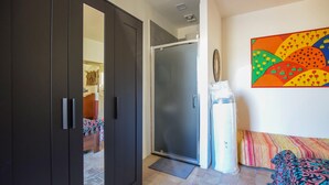 A room with a dark wooden wardrobe, a sliding glass door leading to a bathroom, a colorful abstract painting on the wall, and a patterned sofa.