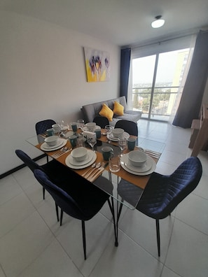A dining area with a glass table set for six, surrounded by dark blue chairs. A grey sofa with yellow cushions is against the wall, and a painting is visible above it.