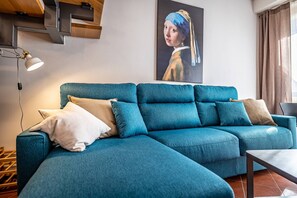 Um sofá azul-esverdeado com várias almofadas, uma luminária e um quadro de uma mulher com um lenço azul na parede.