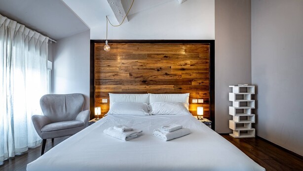 A modern hotel room with a large bed, a wooden headboard, a white chair, bedside lamps, and a white storage unit.