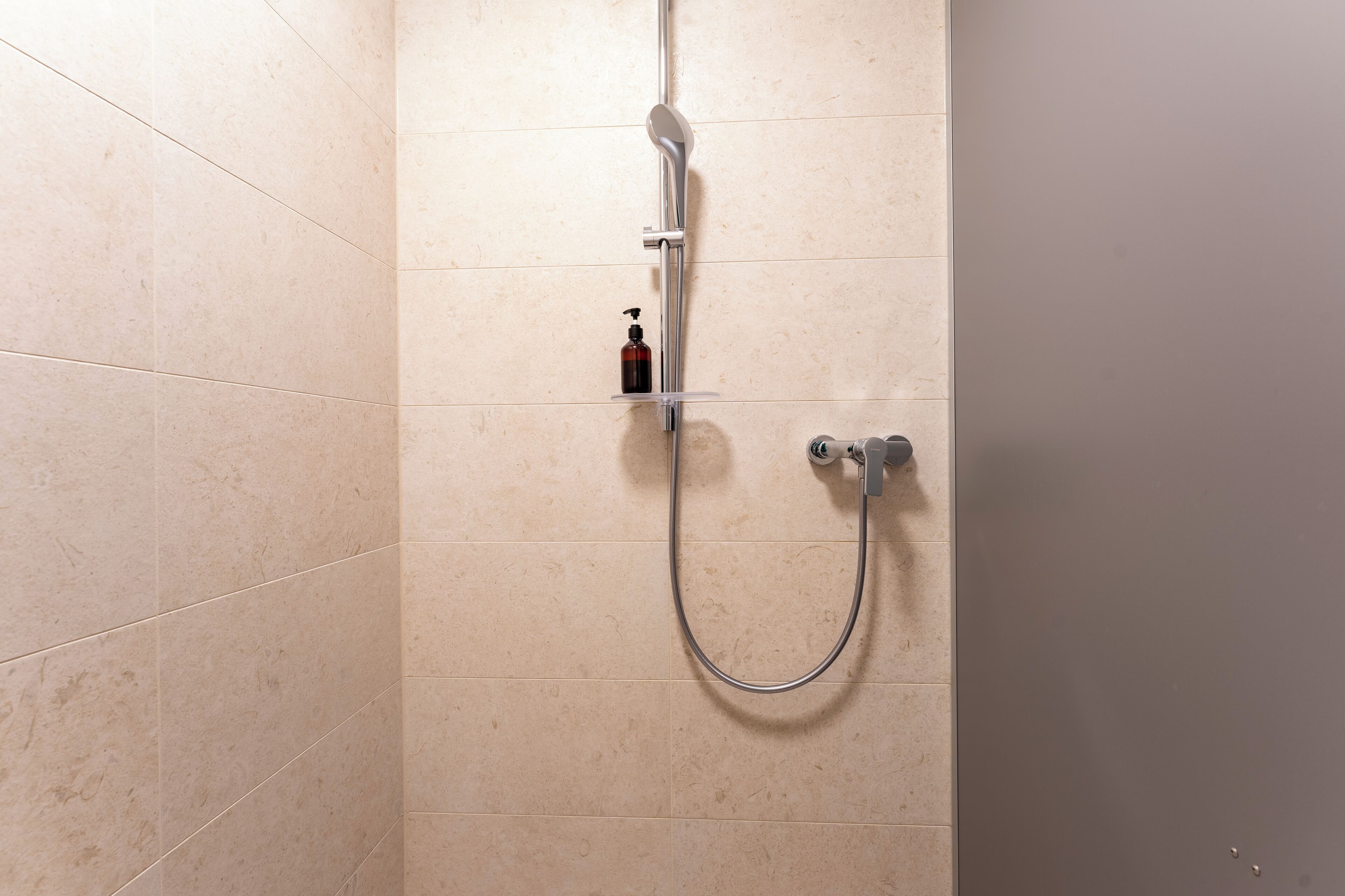A shower with a handheld showerhead and a bottle of shampoo on a shelf.