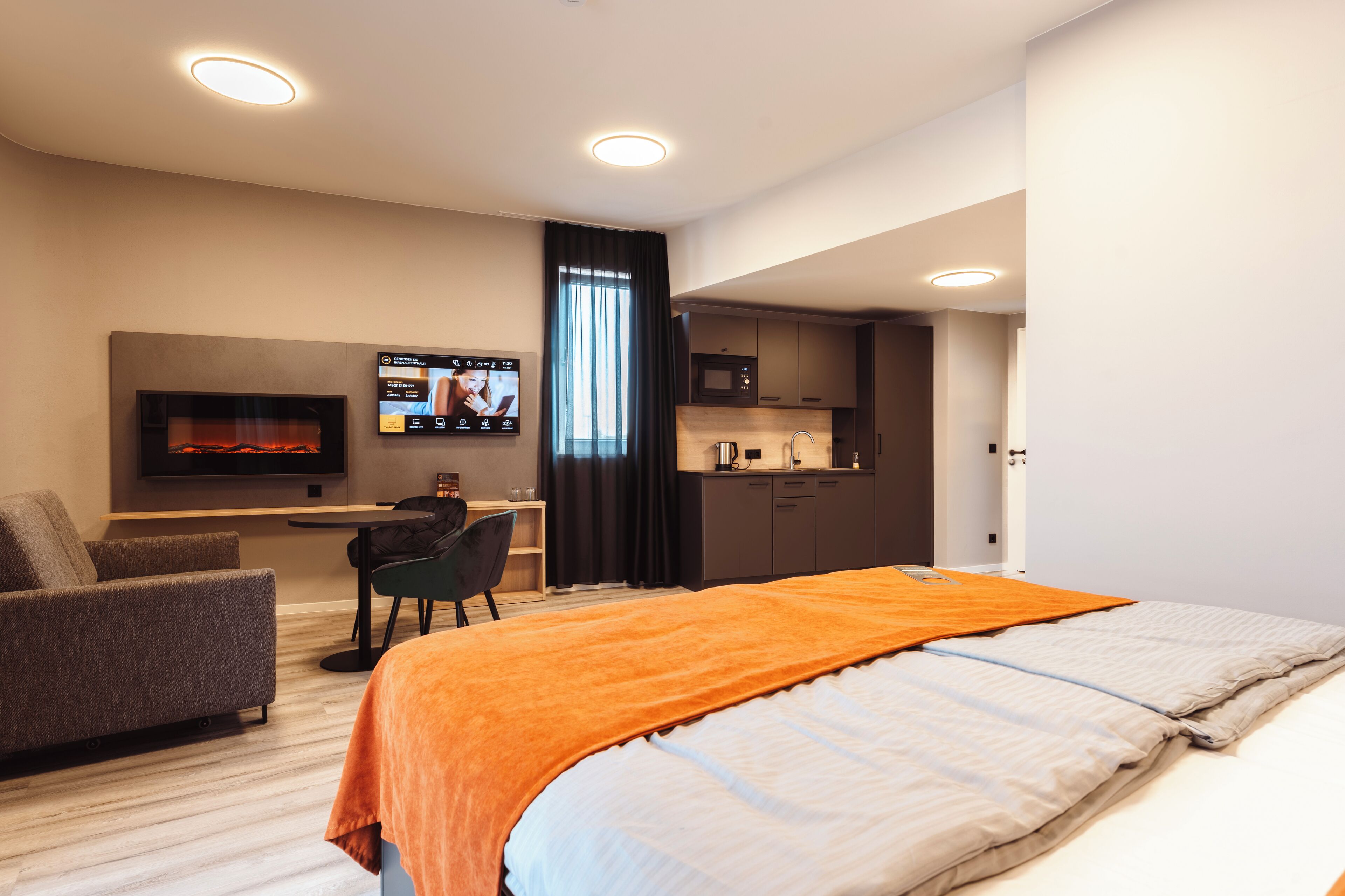 A modern hotel room with a bed, a desk, a TV, and a kitchen area.