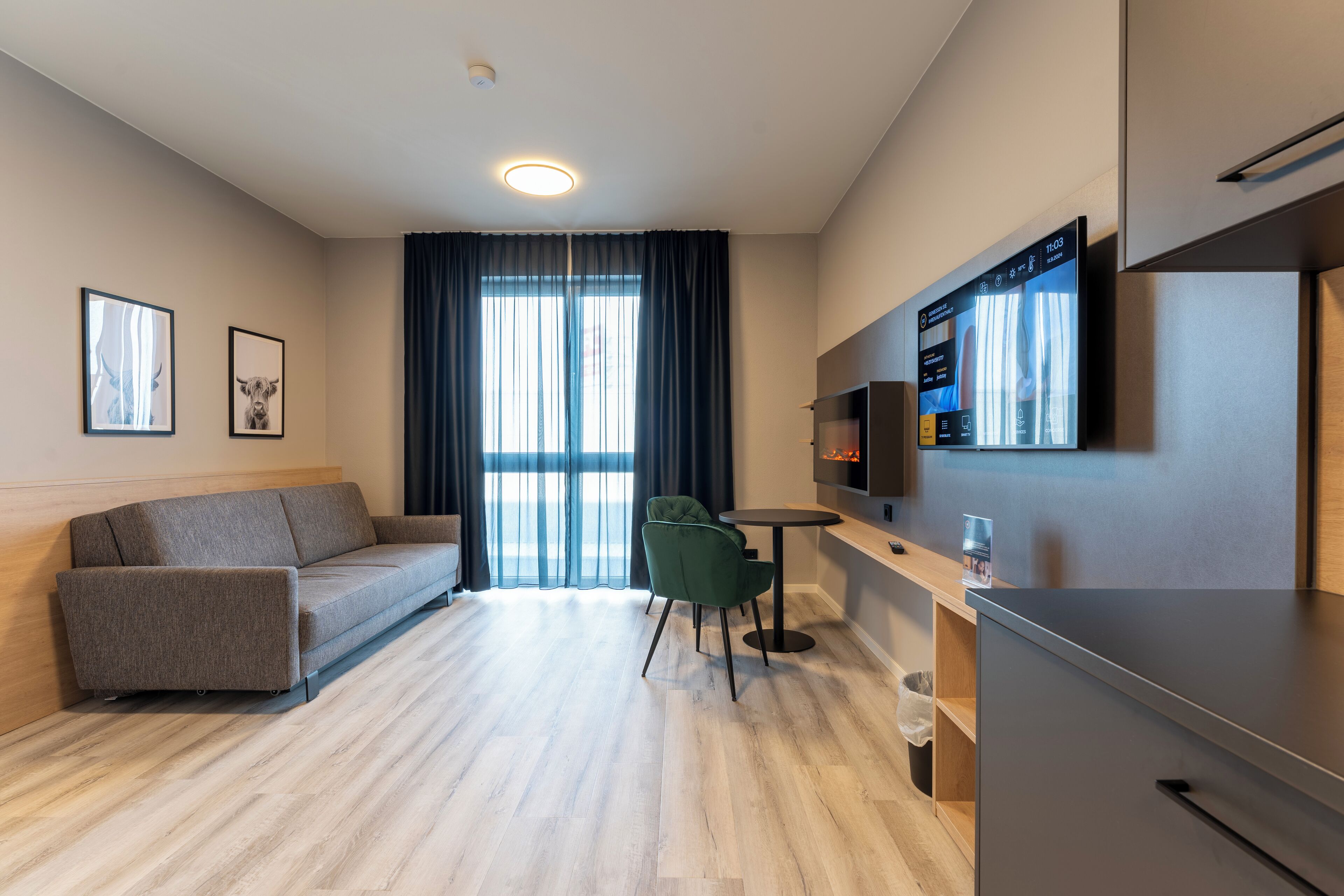 A modern, well-lit room with a sofa, a dining table with a chair, a TV mounted on the wall, and a refrigerator.