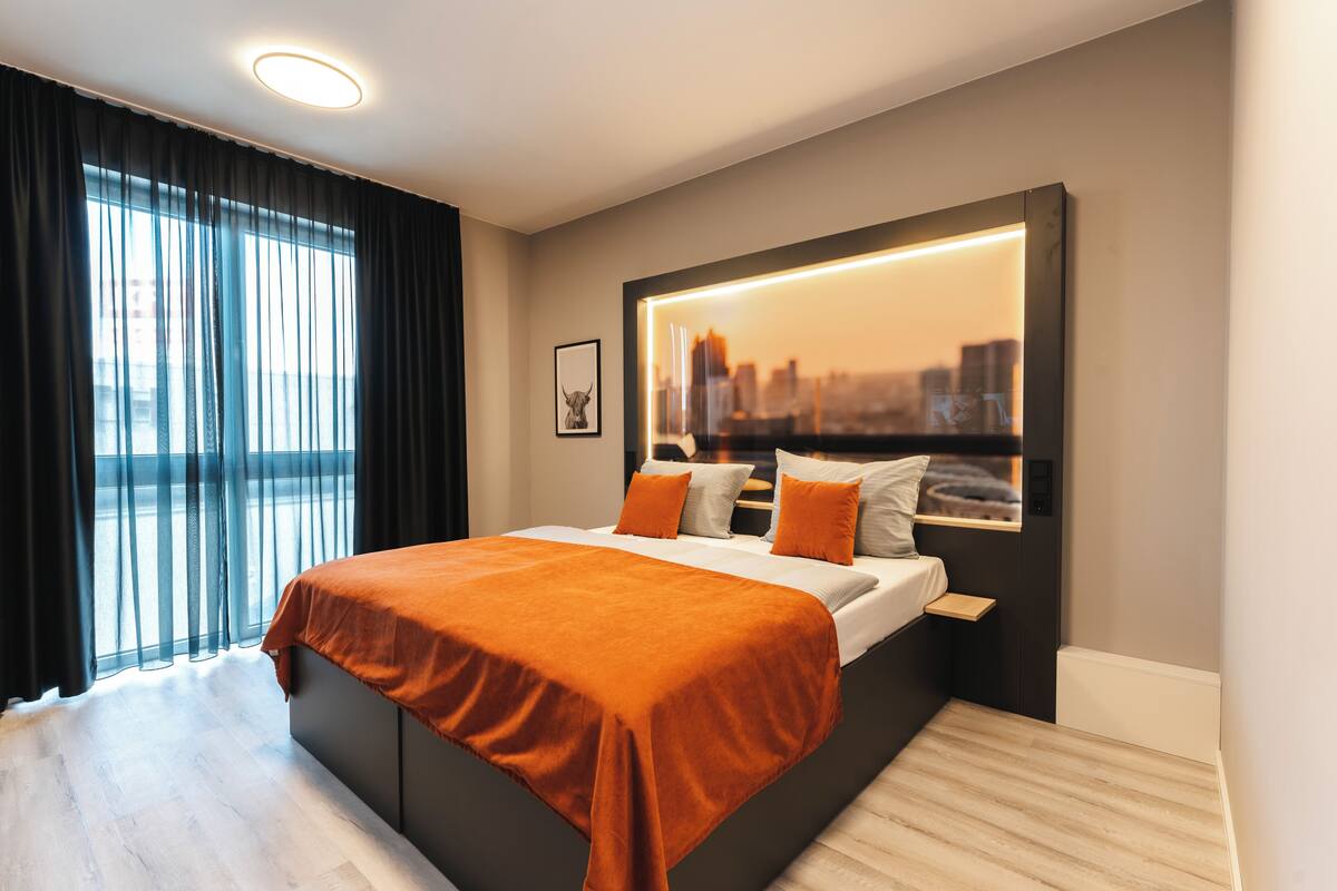 A modern hotel room with a large bed, a city view through a window, and a cityscape artwork on the wall.