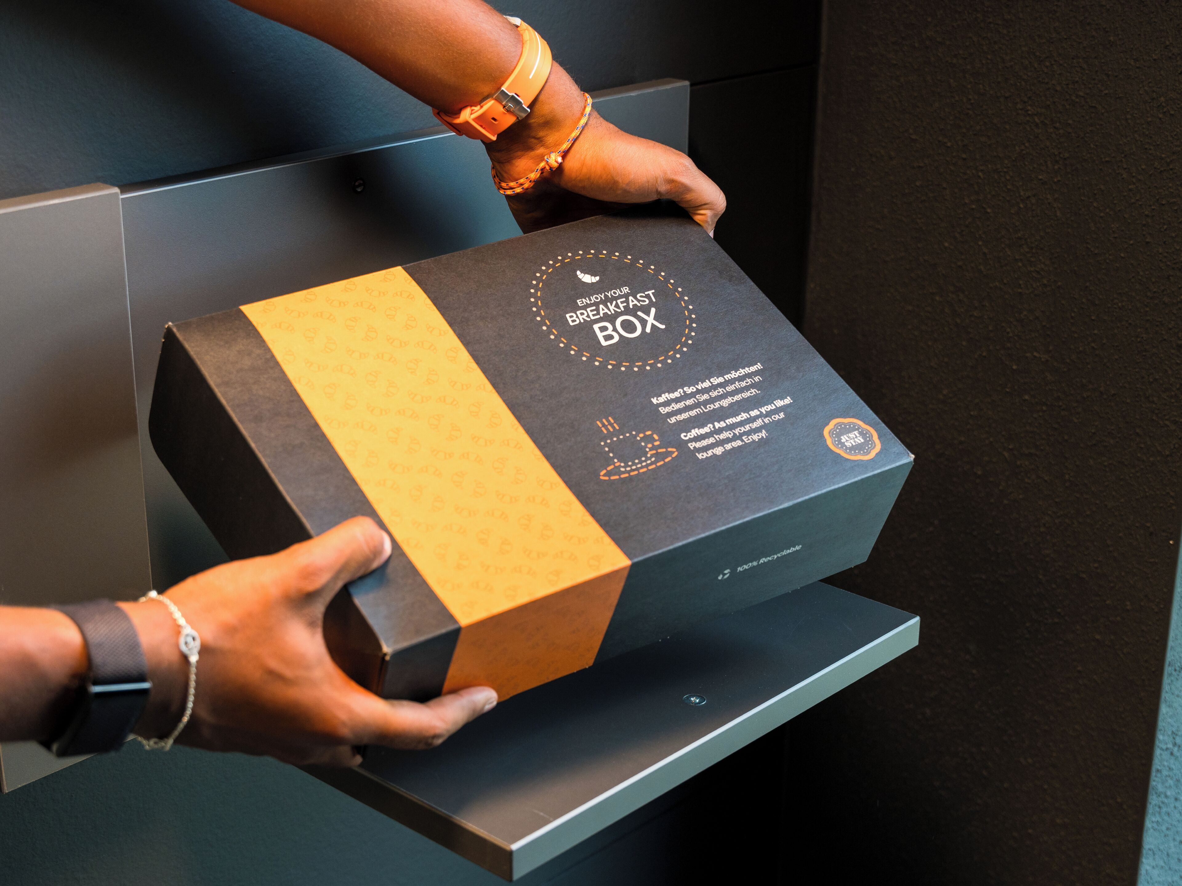 Una persona sosteniendo una caja etiquetada como "ENJOY YOUR BREAKFAST BOX" con un mensaje sobre café y una nota 100% reciclable.