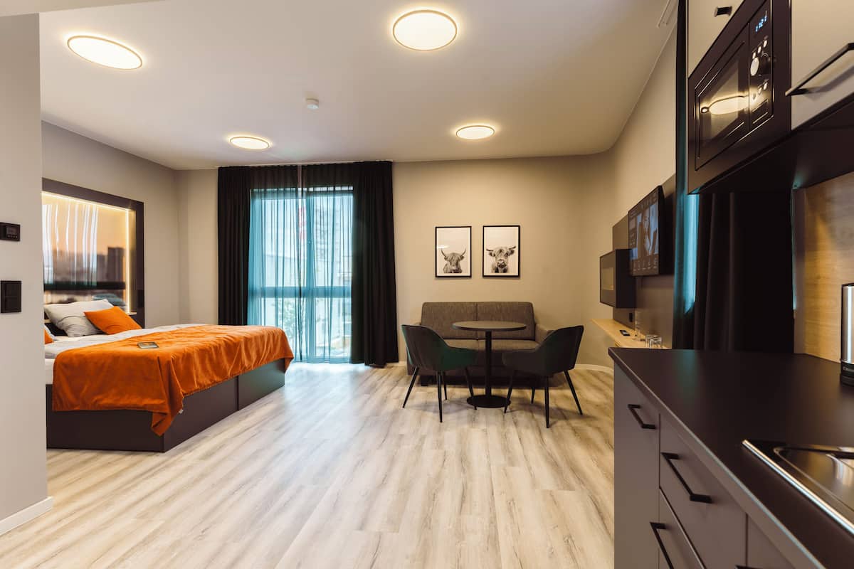 A modern hotel room with a bed, a small dining area, a kitchen, and a large window with curtains.
