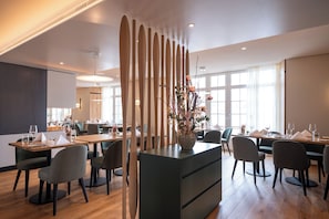 A modern dining area with wooden tables and chairs, a central decorative plant, and large windows allowing natural light. - See & Park Hotel Feldbach (Steckborn)