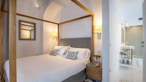 A hotel room with a large bed, two bedside lamps, a wooden headboard, a nightstand with a vase of flowers, and a bathroom with a sink and mirror. - Suite Agave close to the centre of Sorrento (Sant´Agnello)