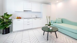 Una cocina moderna con armarios blancos, un fregadero y un bol de frutas. Hay una mesa verde con un jarrón blanco y una orquídea. Un sofá azul claro está apoyado en la pared.