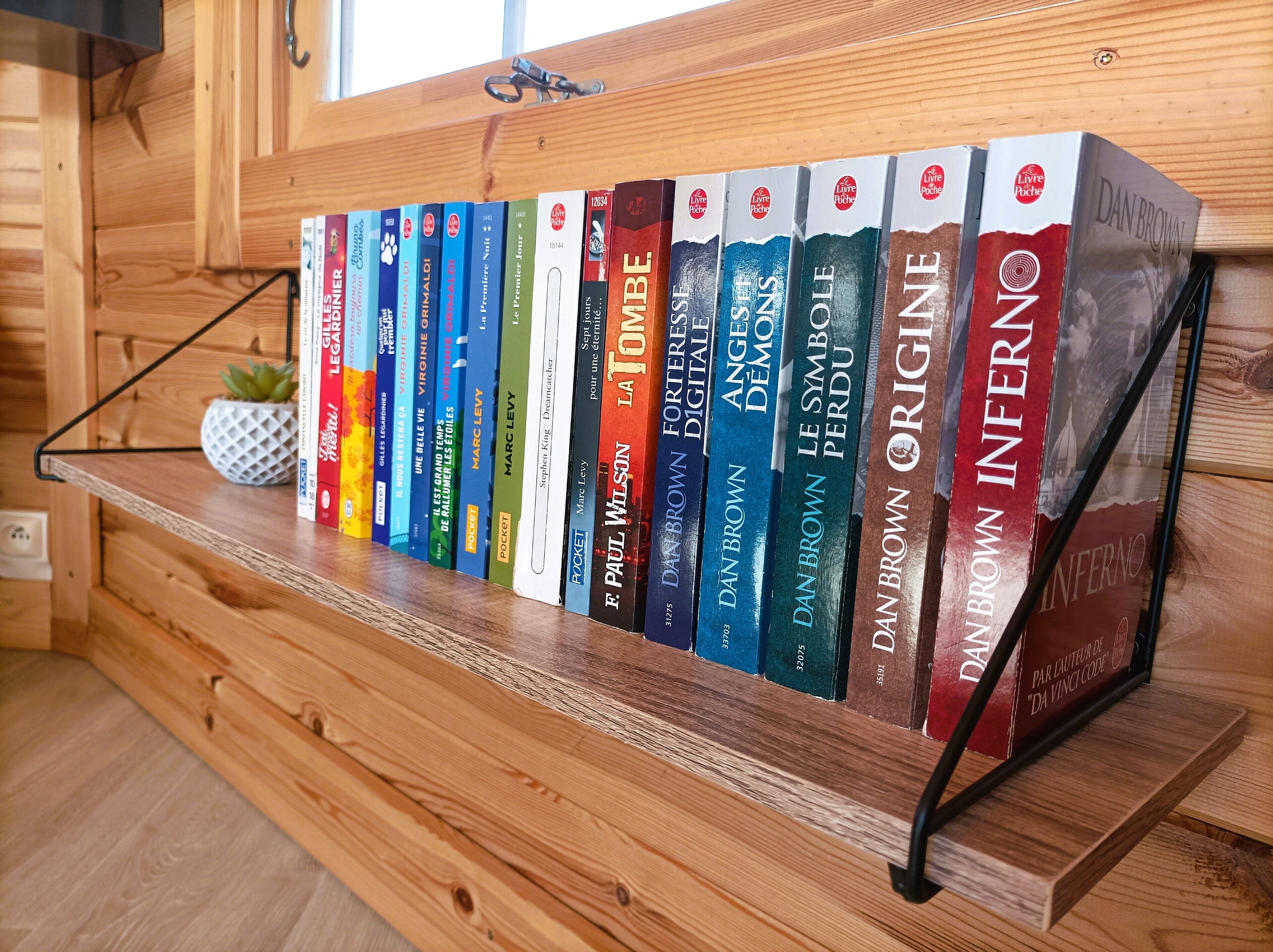 A wooden shelf with a row of books, including titles by Dan Brown, and a small potted plant.