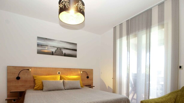 A bedroom with a wooden headboard, a bed with striped bedding, two bedside lamps, a wall-mounted picture, and a green armchair. - Holiday apartment with air conditioning (Štinjan)