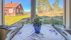 A room with a table set for two, a vase with flowers, and a view of a red house and trees through large windows. - Cozy home in Strömstad with WiFi (Strömstad)