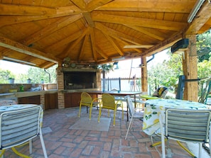 An outdoor kitchen area with a brick floor, wooden ceiling, and a brick fireplace. There are yellow chairs and a table with a tablecloth. - Holiday apartment with air conditioning (Medulin)