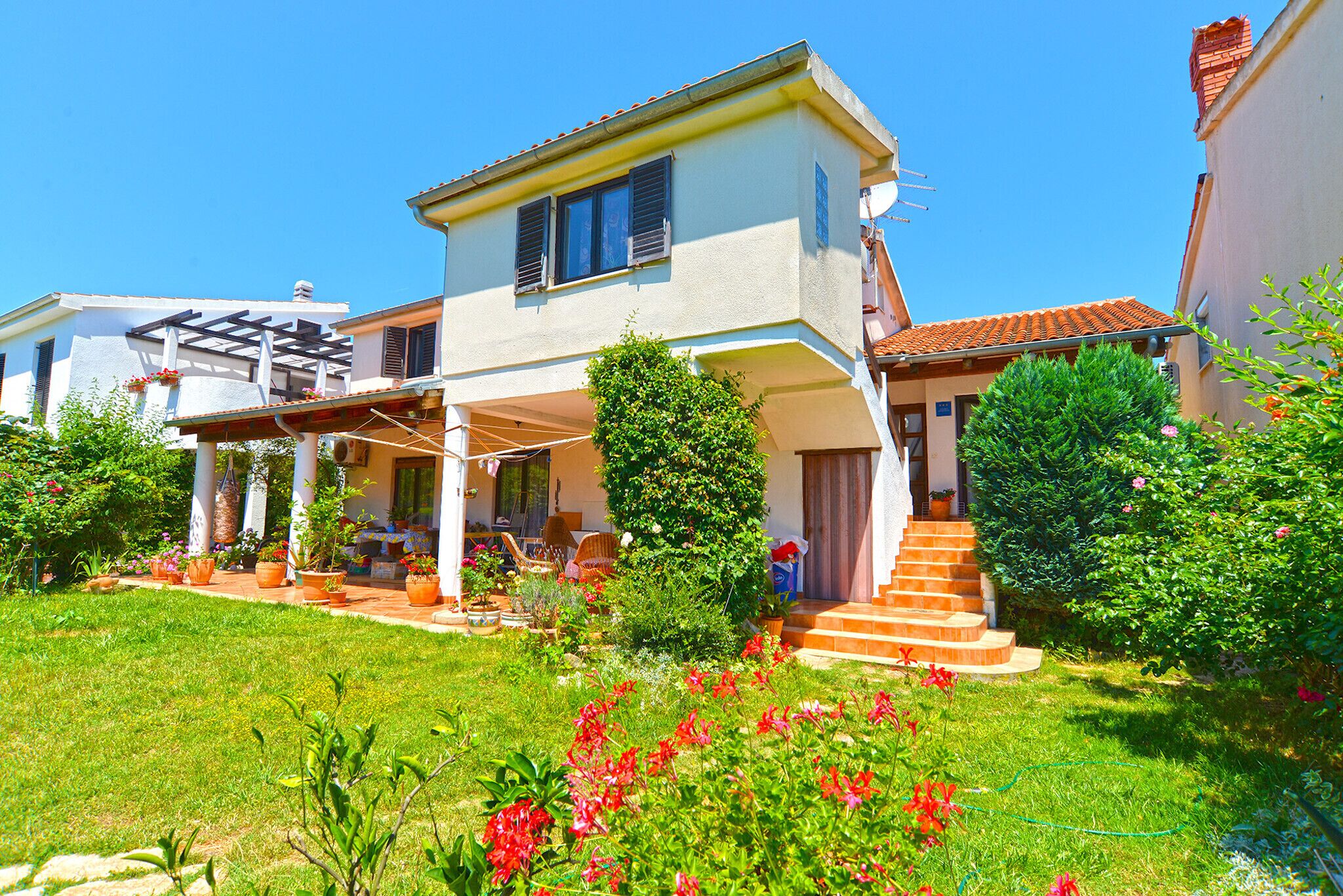 A two-story house with a covered patio, a garden with various plants and flowers, and a staircase leading to the entrance.