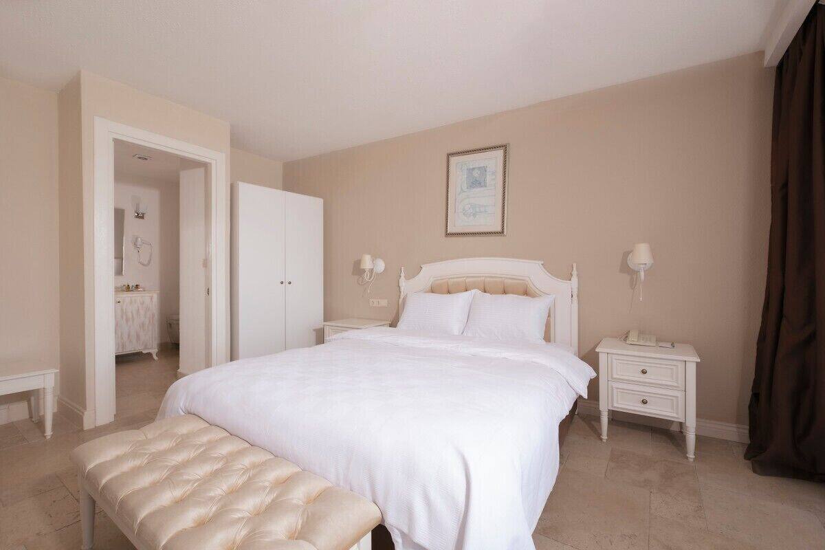 Een slaapkamer met een groot bed, nachtkastjes, een bankje en een deur naar een andere kamer.