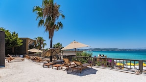 Un salon de plage doté de chaises longues en bois et de parasols, offrant une vue sur une mer d’un bleu limpide et sur une côte lointaine.