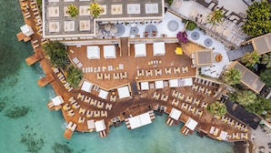 Vue aérienne d’un complexe hôtelier doté de plusieurs terrasses, certaines aménagées avec des chaises longues et des parasols, et d’autres avec des tables et des chaises.