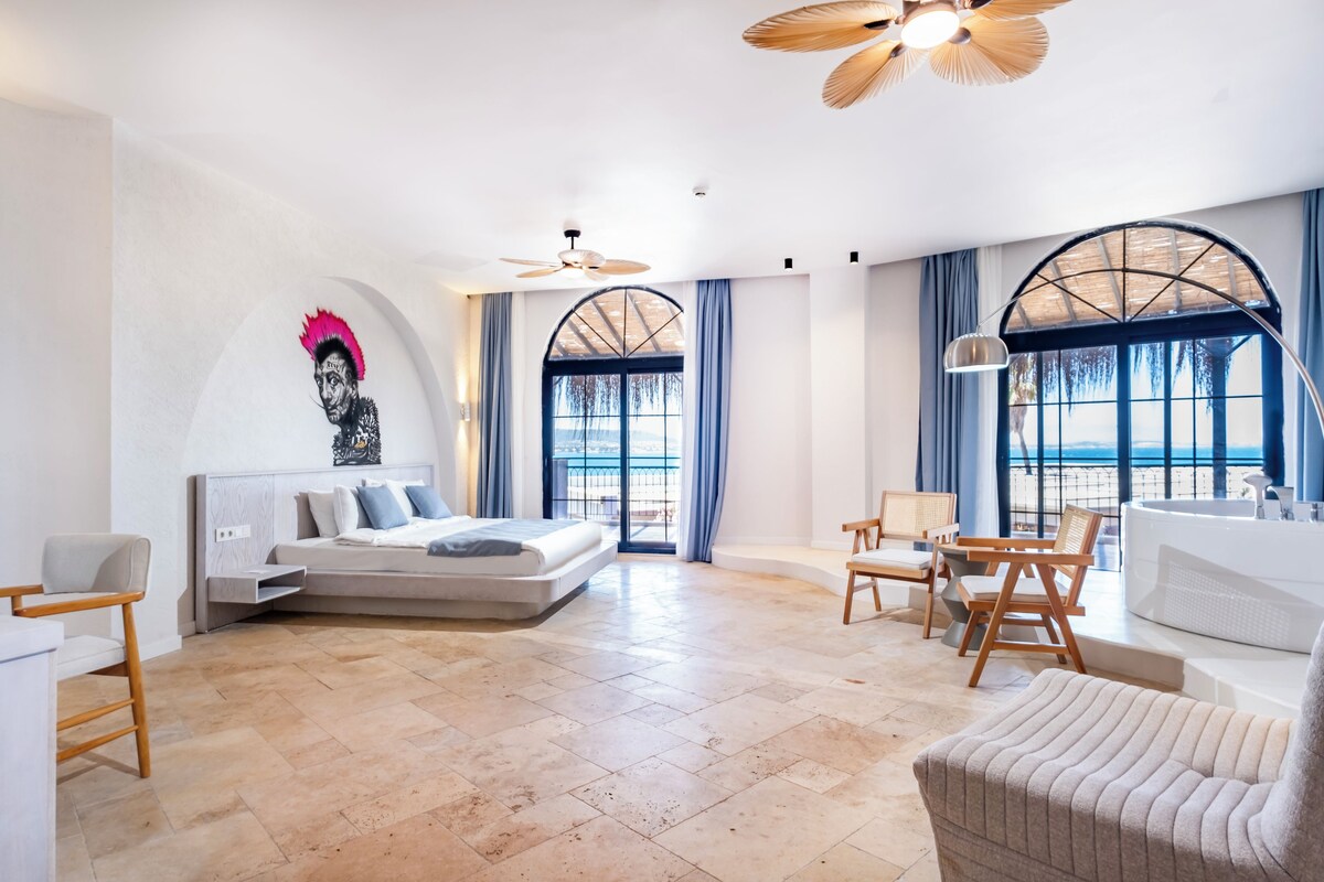 Una habitación de hotel moderna con cama, zona de estar y ventanas amplias con vista al mar.