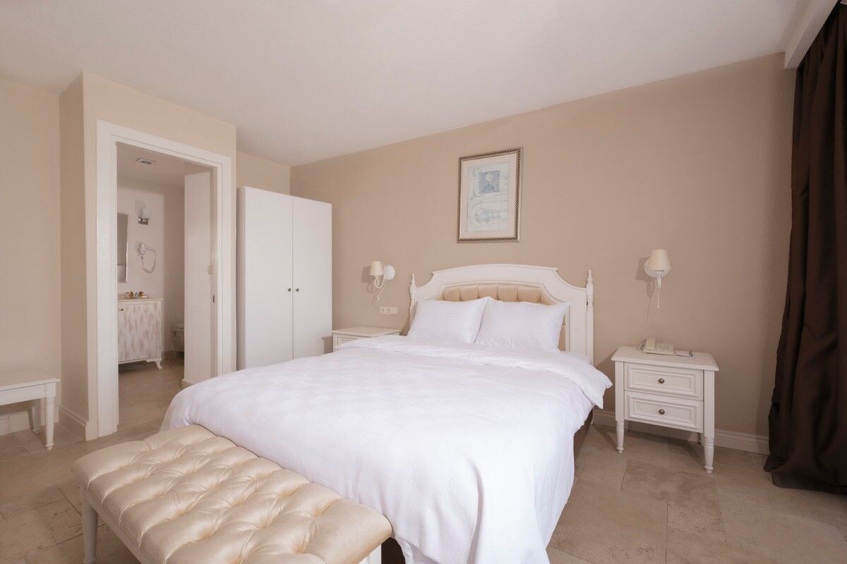 A bedroom with a large bed, bedside tables, a bench, and a door leading to another room.