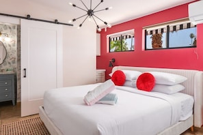 A bedroom with a bed, white bedding, red pillows, a ceiling light fixture, and a window with a view of greenery.