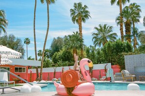 Een zwembad met een flamingo opblaasbaar speeltje, palmbomen en een rood gebouw met een witte luifel.