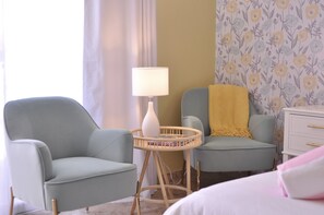 A room with two light blue armchairs, a small round table with a white lamp, and a white dresser with gold handles.
