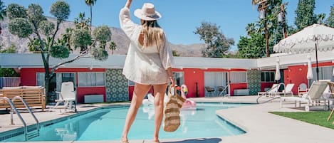 Un área de piscina con sillones reclinables, una piscina y un edificio con paredes exteriores rojas y blancas.