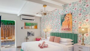 A bedroom with a green upholstered headboard, a large bed with white bedding, two bedside tables with lamps, a floral wallpaper, and a poster on the wall.