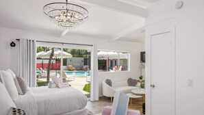 A modern hotel room with a large bed, a sofa, and a small table. There is a balcony with a pool and umbrellas.