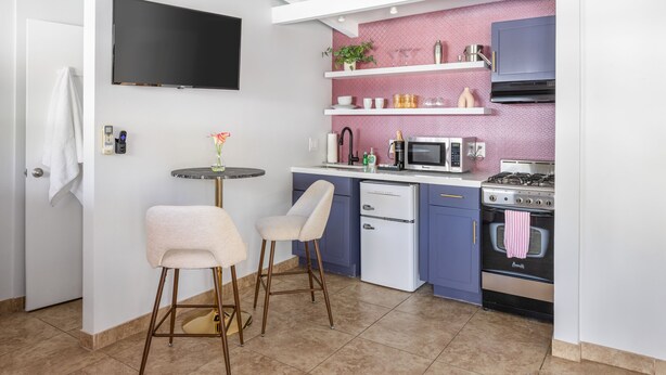 A compact kitchen with a flat-screen TV, a microwave, a stove, and a sink. There are two bar stools and a small table with a vase of flowers. - The Sofia Suite · Adults-Only Palm Springs Pool (Palm Springs)