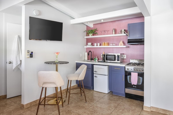 A compact kitchen with a flat-screen TV, a microwave, a stove, and a sink. There are two bar stools and a small table with a vase of flowers.