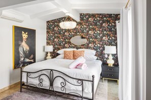 A bedroom with a floral wallpaper, a bed with white linens, a black metal headboard, a nightstand with a lamp, and a painting on the wall.