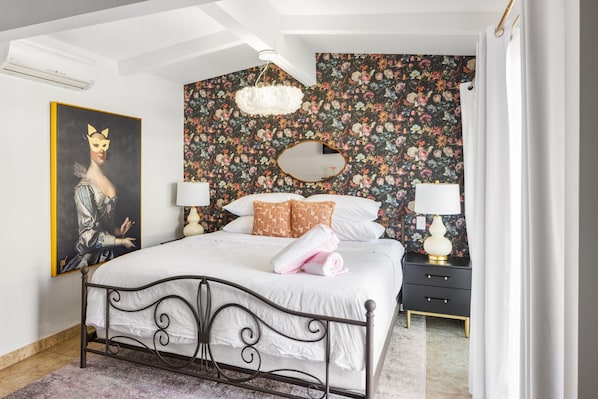 Ein Schlafzimmer mit floralem Wandpapier, einem Bett mit weißen Bettwäsche, einem schwarzen Metall-Kopfstück, einem Nachttisch mit Lampe und einem Gemälde an der Wand.