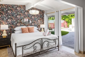 Ein Schlafzimmer mit Blumen-Tapete, einem Bett mit weißer Bettwäsche, einem Spiegel und Blick auf einen Garten durch eine offene Tür.