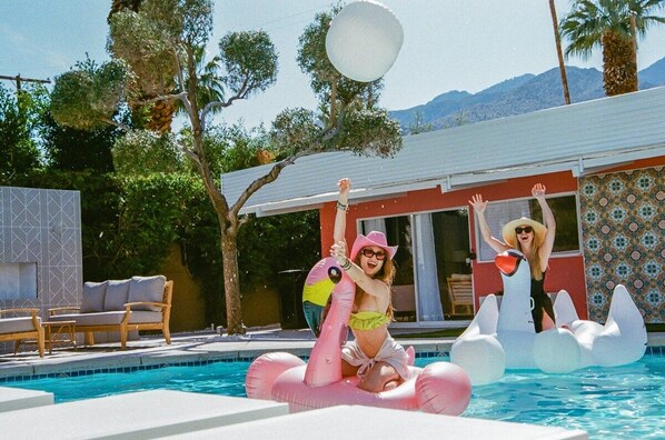 A swimming pool with inflatable flamingo floats, a lounge chair, and a building with patterned tile walls.