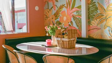 A dining area with a round table, wicker chairs, a vase with flowers, and a wall mural.