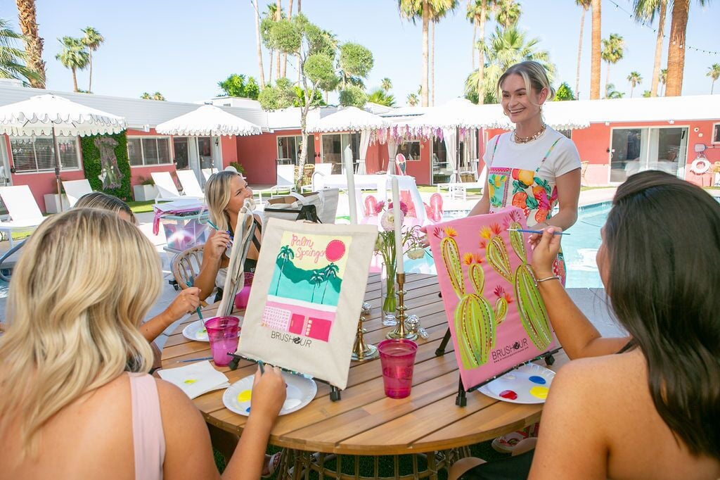 A group of people painting at a wooden table with beach-themed artwork and a 'Brush Hour' tote bag.