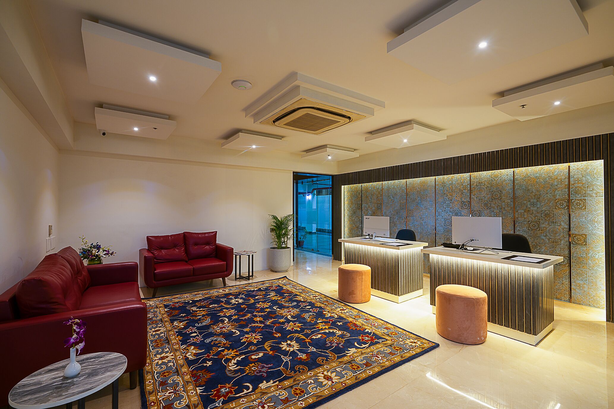 Un hall d’hôtel avec un comptoir d’accueil, deux canapés rouges, un grand tapis à motifs et une plante en pot.