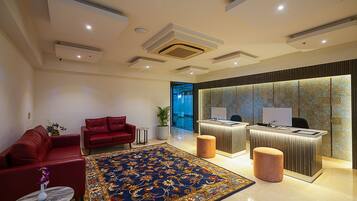 A hotel lobby with a reception desk, two red sofas, a large patterned rug, and a potted plant.