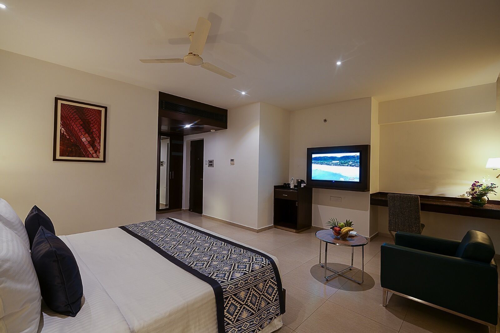 A hotel room with a bed, a desk with a TV, a chair, and a small table with fruit.