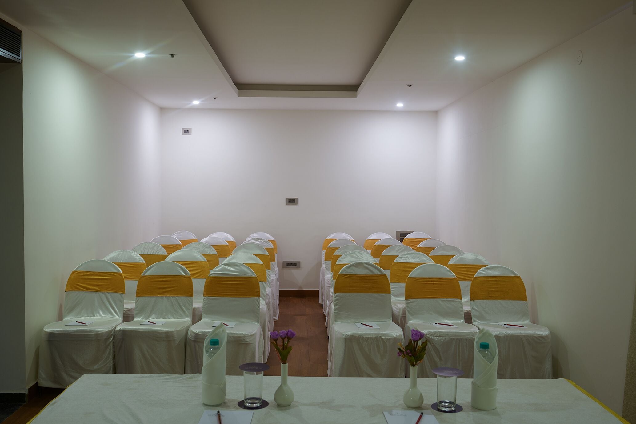 Un salón de conferencias con filas de sillas, cada una con una funda blanca y cintas amarillas. Hay jarrones con flores sobre la mesa.