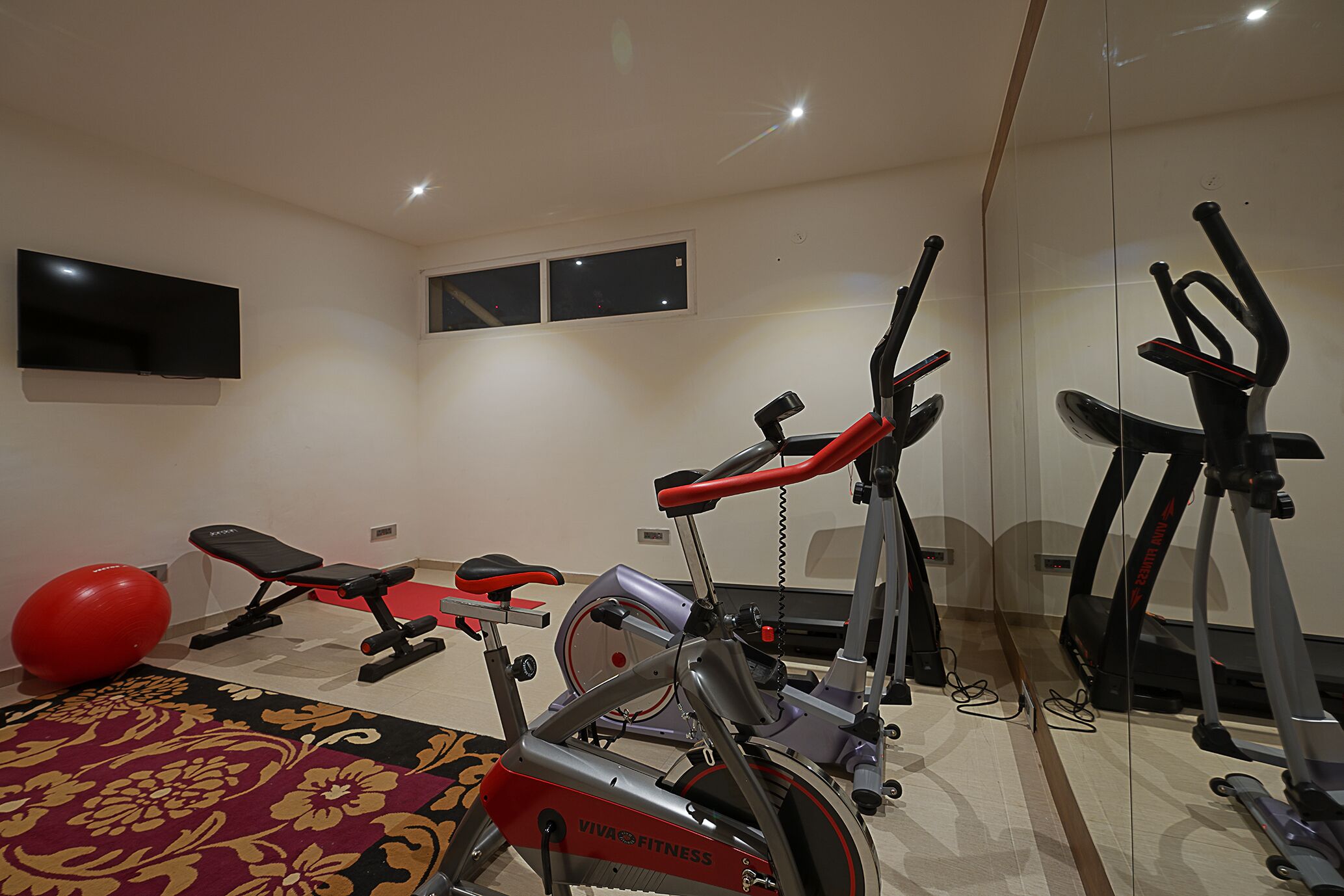 Uma academia em casa com equipamentos de ginástica, incluindo uma bicicleta ergométrica, elíptico e uma bola de exercício vermelha.