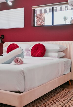 A bed with white bedding and red pillows, a window with a view of palm trees, and a red wall. - The Edie Suite · Palm Springs Pool & Hot Tub (Palm Springs)