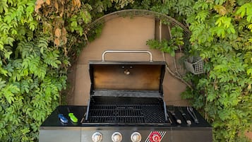 A black outdoor grill with a metal handle and two visible control knobs, surrounded by dense green foliage.