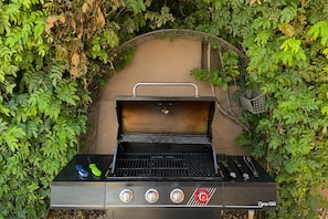 A black outdoor grill with a metal handle and two visible control knobs, surrounded by dense green foliage. - Bowie Suite · Palm Springs Pool, Hot Tub, Kitchen (Palm Springs)